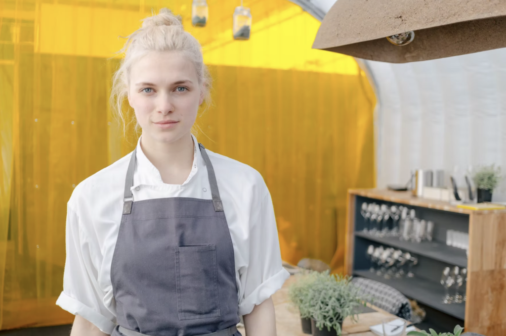 Laura Paine chef with blonde hair. Wearing grey apron and standing in kitchen with yellow backdrop.