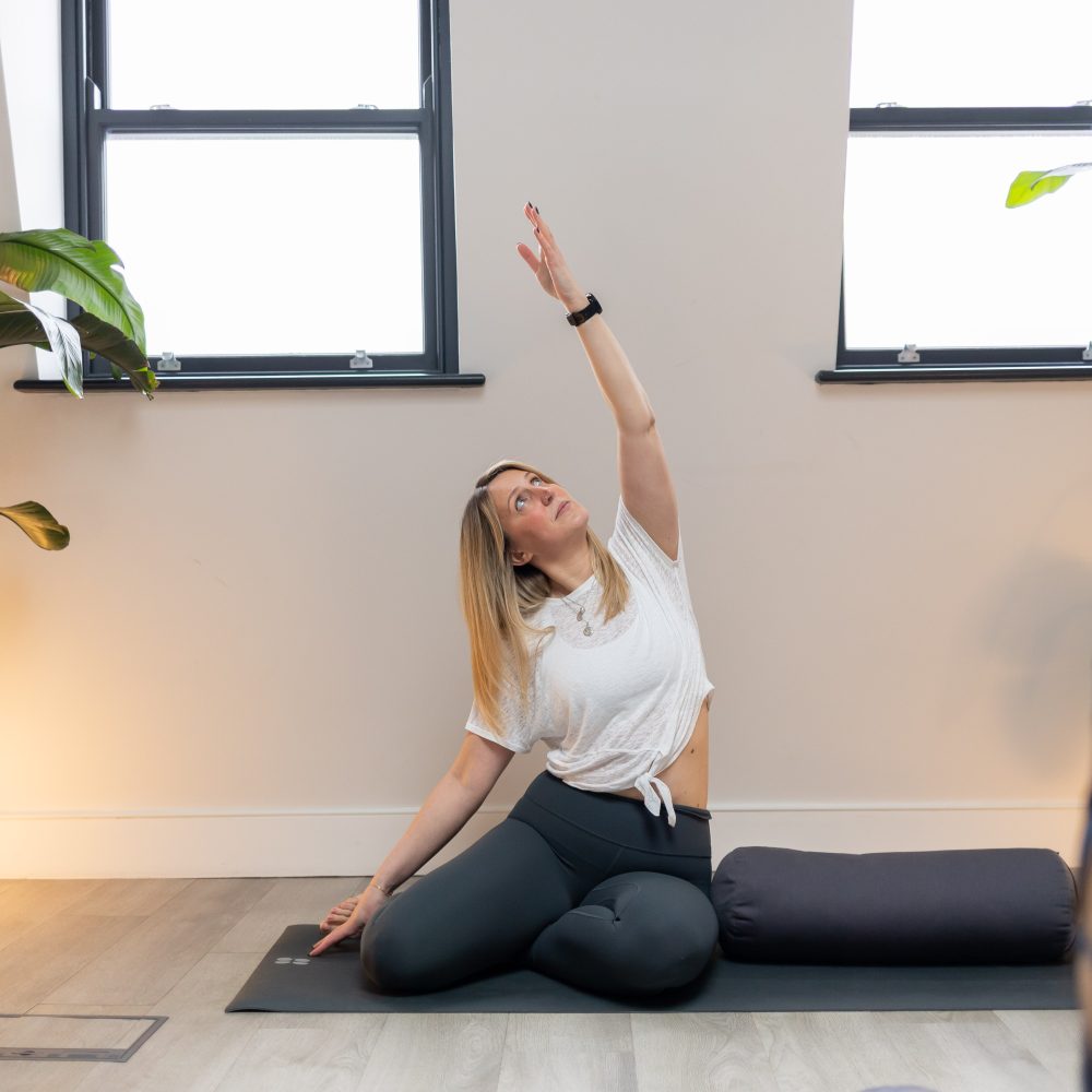 Martha sat on a mat with legs to one side and left arm lifted. she is sat next to a bolster in front of 2 windows.