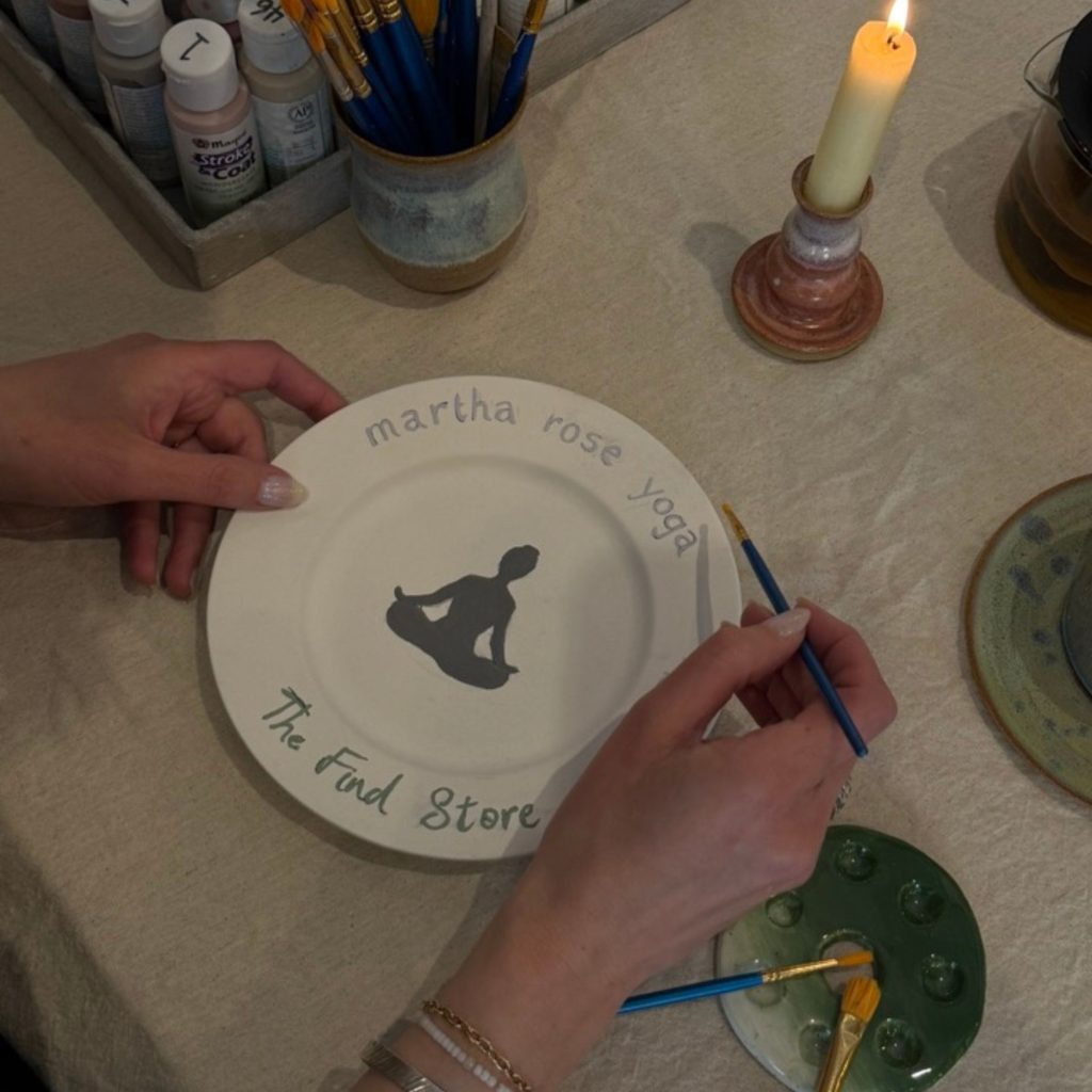 Martha painting a small plate with the Martha Rose Yoga and The find Store branding and a silhouette of someone sat in lotus pose in the centre. On the table are paints, paint brushes and a candle in candlestick holder