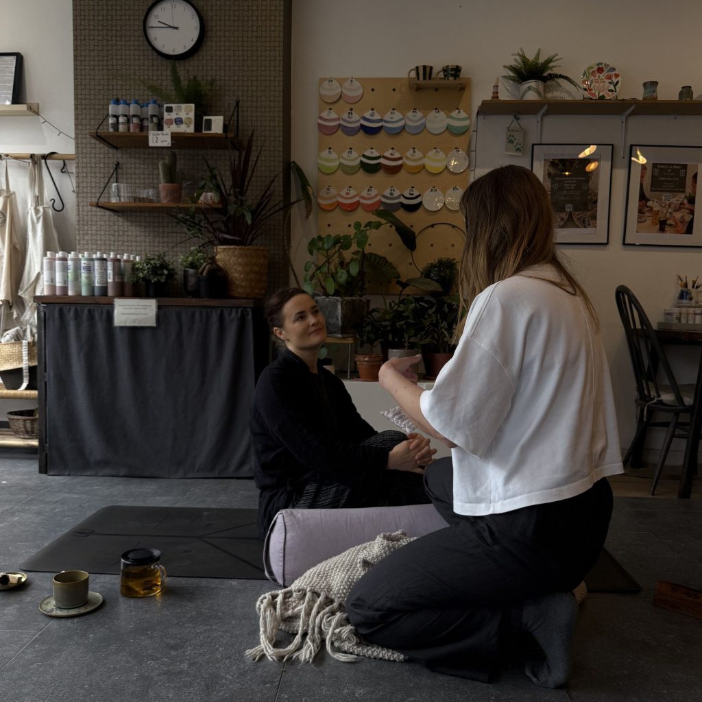 Sev and Martha talking in the find store surrounded by ceramics, paints, plants and more. Martha holds a lavender eye pillow. There is a bolster, blanket, pot of tea and a cup on the floor